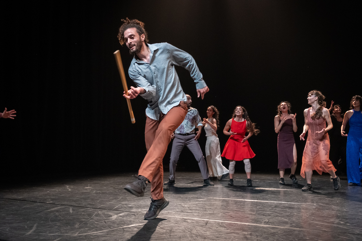 Lead dancer with stick as ensemble in colorful dresses clap and cheer during Badke Remix performance
