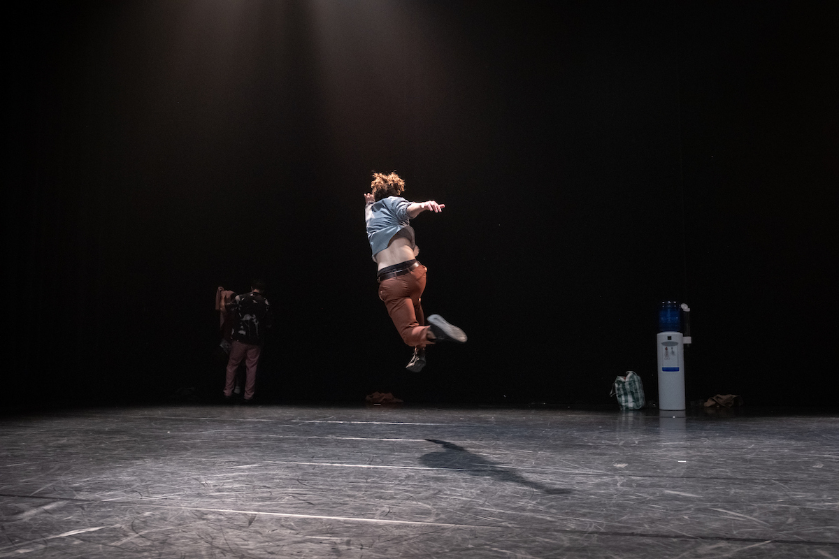 Dancer mid-air in a dramatic jump during Badke Remix performance, wearing blue shirt and orange pants