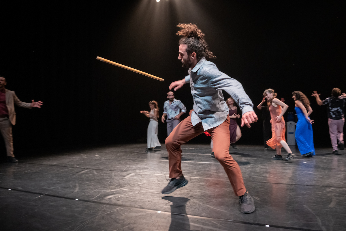 Lead dancer performing with traditional stick prop while ensemble dancers move in background during Badke Remix